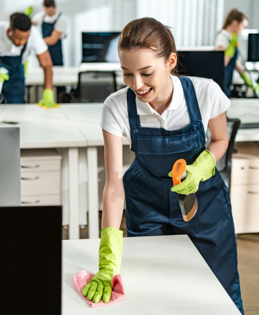 cheerful cleaner in overalls cleaning office desk with rag